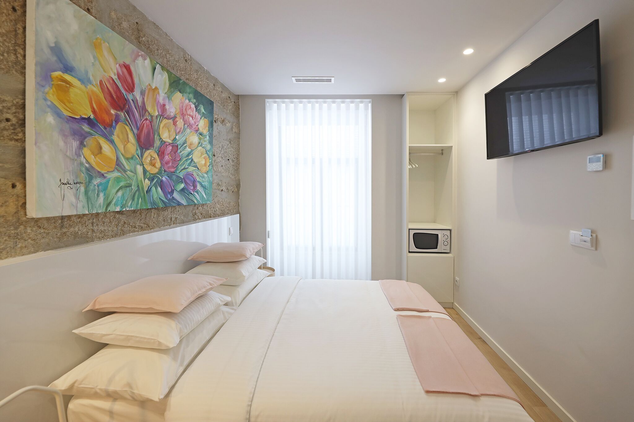 A bedroom with a bed, pillows, a painting, a TV, and a window with blinds.