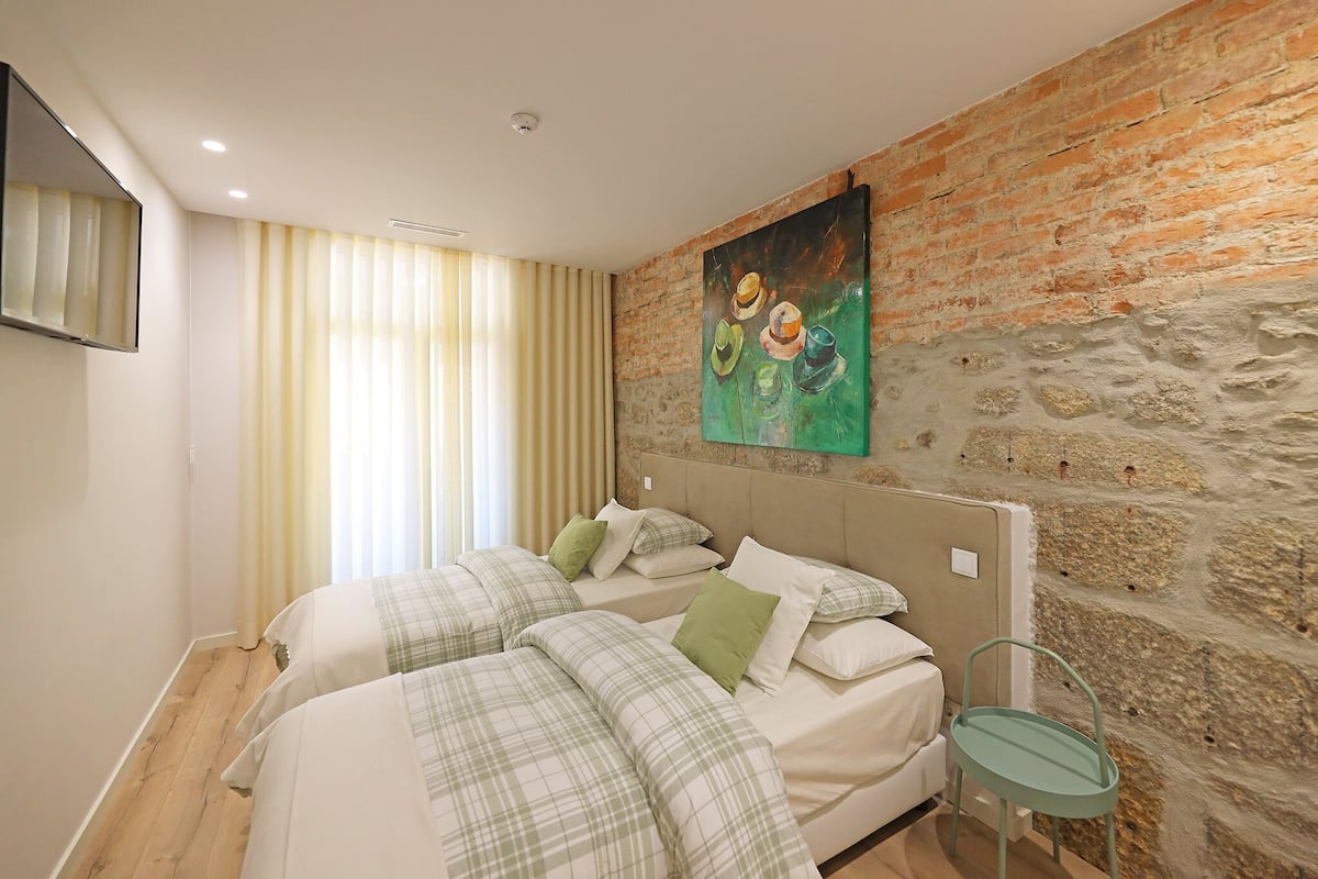 A bedroom with two beds, a stone wall, a painting, and a window with curtains.