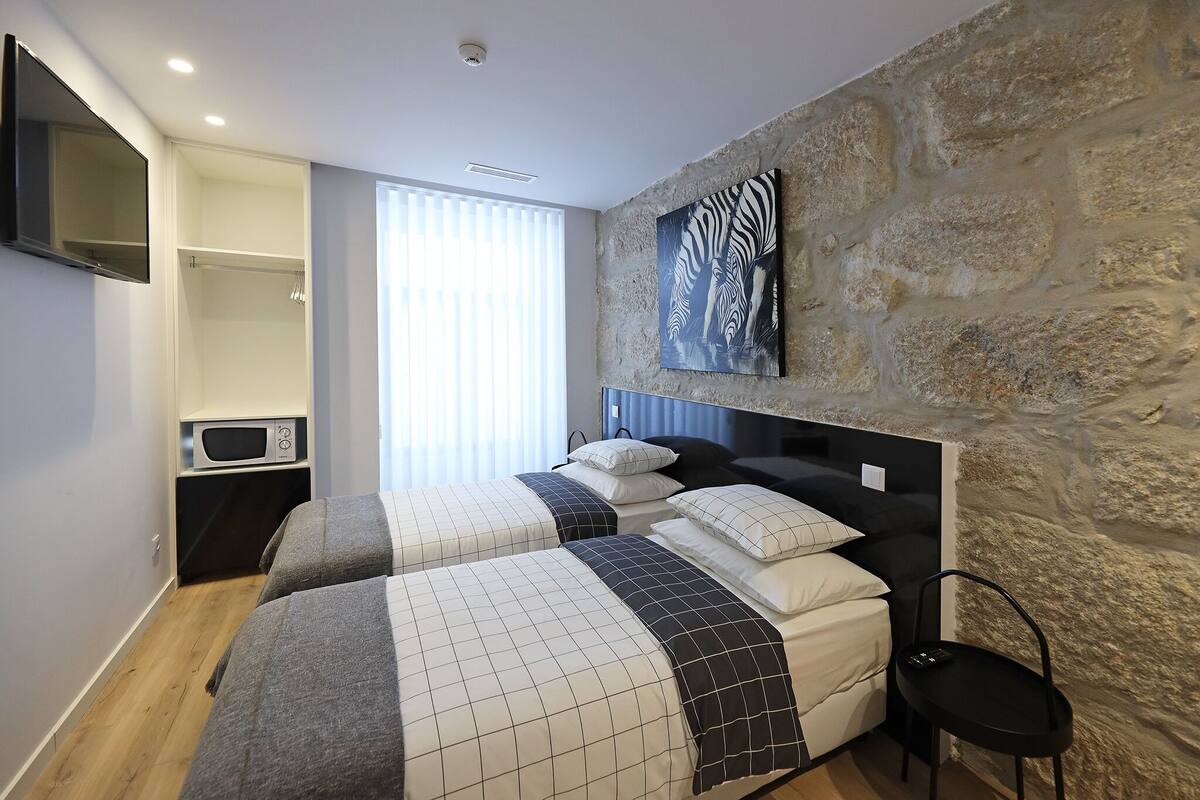 A bedroom with two beds, a stone wall, a mounted TV, and a window with blinds.
