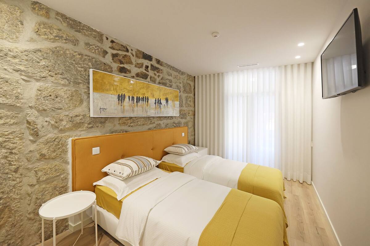 A hotel room with two beds, a stone wall, a painting, a TV, and a window with curtains.