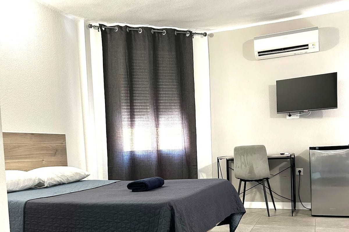 A hotel room with a bed, a desk, a chair, a television, and an air conditioning unit.