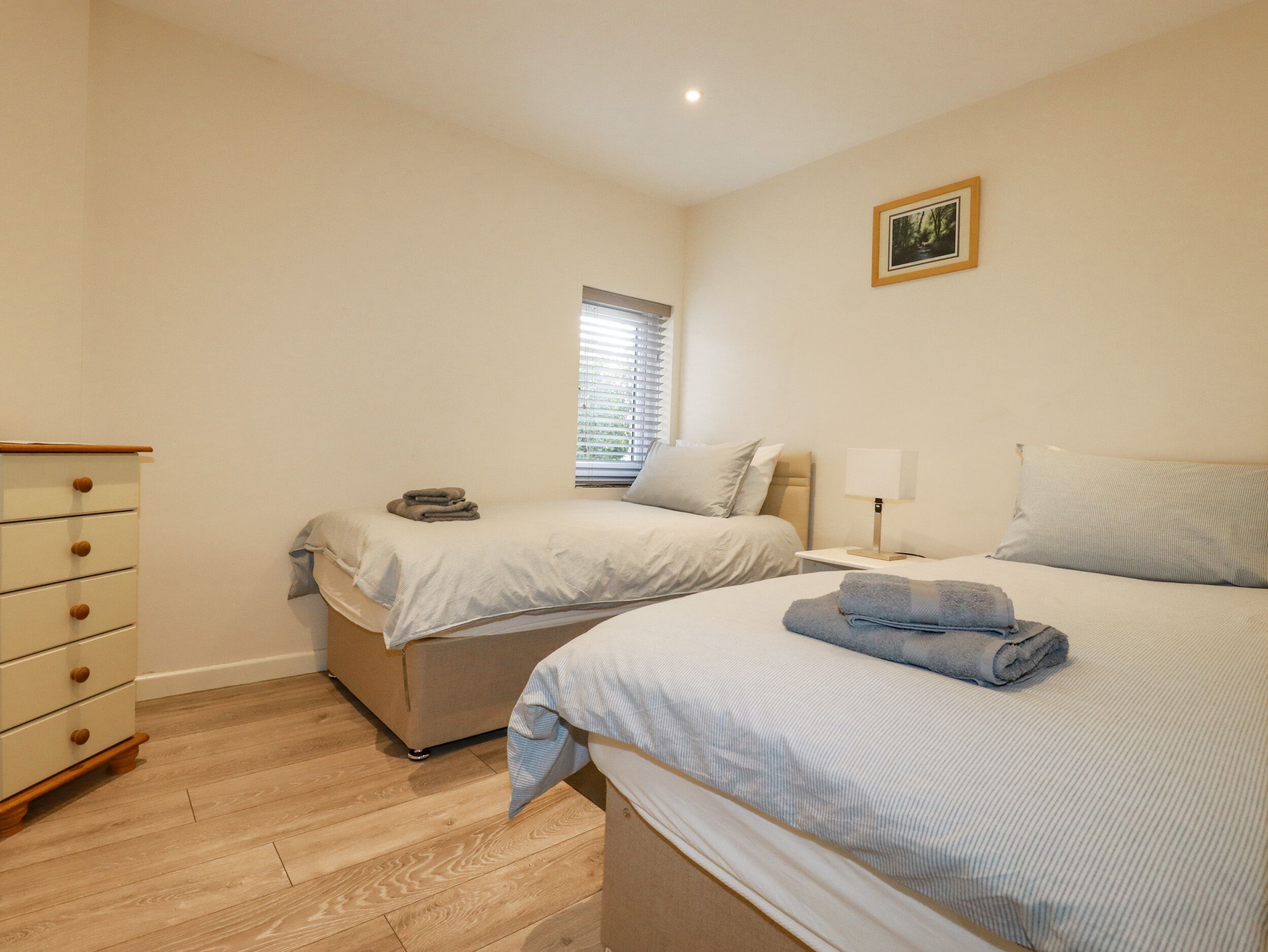 A bedroom with two beds, a dresser, a bedside table with a lamp, and a window with blinds.