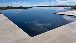 Uma piscina infinita retangular com borda azul translúcida, rodeada por um deck revestido de azulejos.