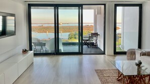 Sala de estar moderna com uma ampla porta de vidro deslizante que dá para um terraço ao ar livre com vista para um corpo d'água e vegetação.