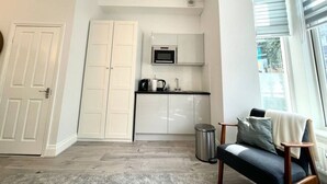A compact kitchen with white cabinets, a microwave, and a coffee maker. There is a trash bin and a chair with a blanket. - F69 21 · London Studios Shepherds Bush near Central Line (London)