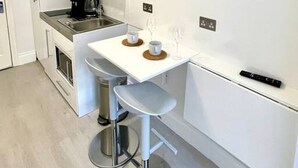 Une cuisine compacte avec des meubles blancs, une petite table à manger, deux tabourets de bar et une machine à café.