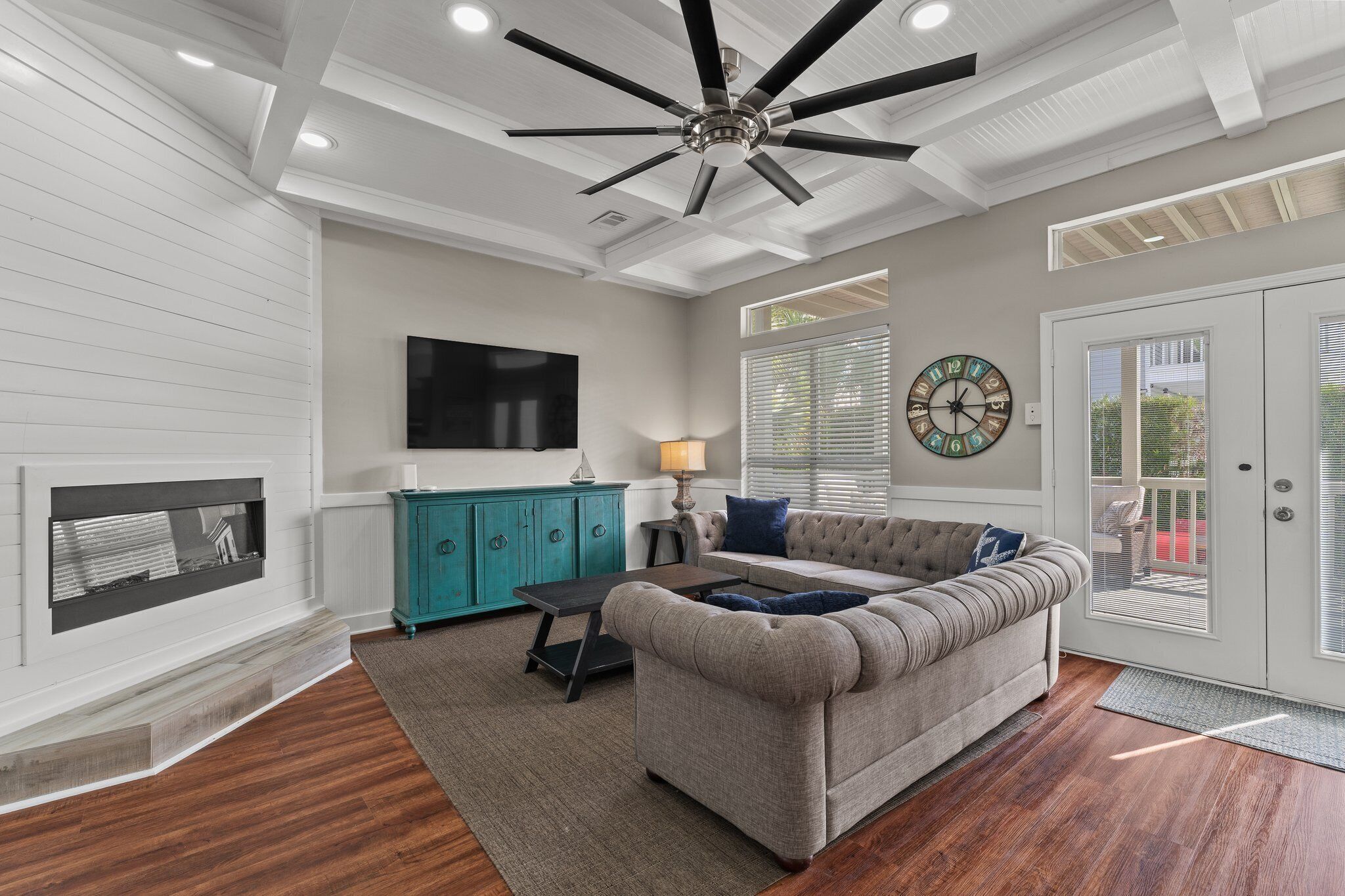 A living room with a fireplace, a large flat-screen TV, a teal cabinet, a gray sofa, and a ceiling fan.