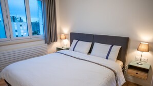 Una cama bien hecha con edredón blanco y dos almohadas, flanqueada por dos mesitas de noche con lámparas, en una habitación con suelo de madera y una ventana con cortinas.