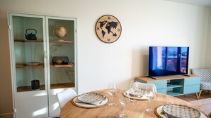 Un comedor con una mesa redonda de madera dispuesta para cuatro personas, un reloj de pared tipo globo y un televisor sobre un mueble azul.