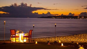 A table set for two with candles on the sand, overlooking a calm sea at sunset.