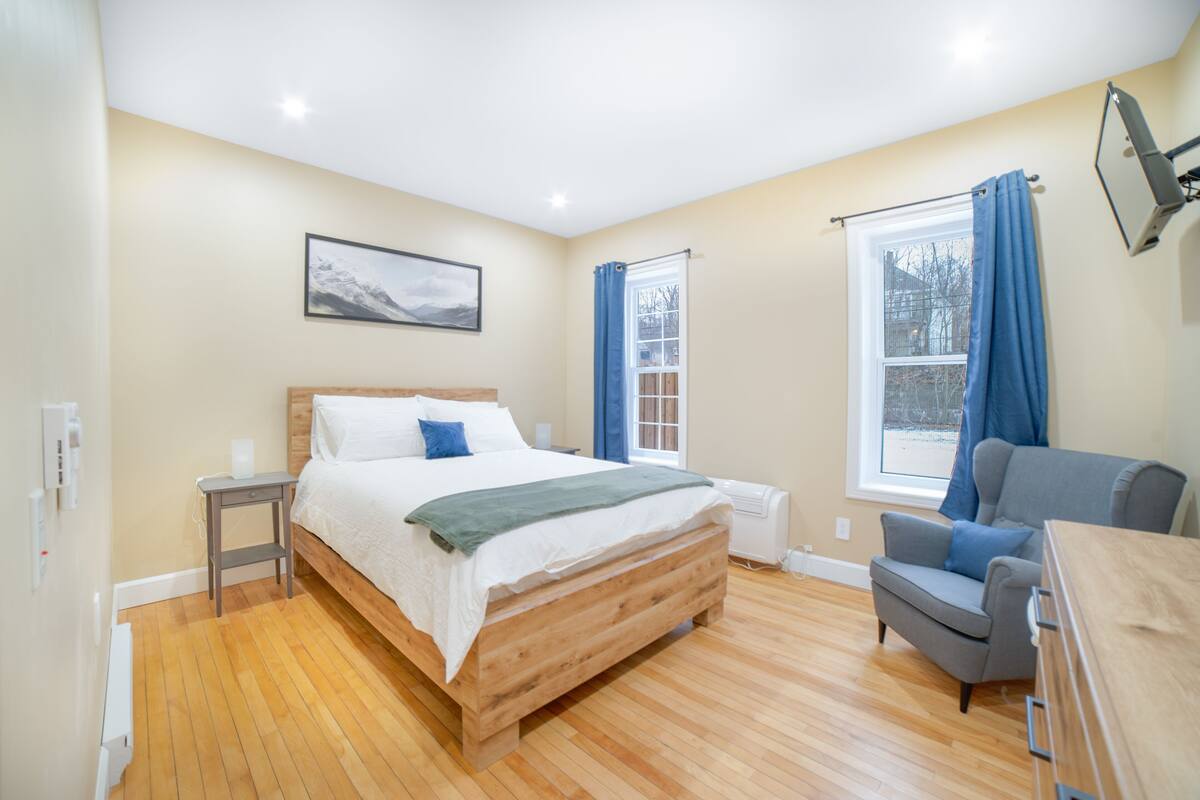 Un dormitorio con una cama de madera, un sillón gris, una ventana con cortinas azules y un televisor montado en la pared.