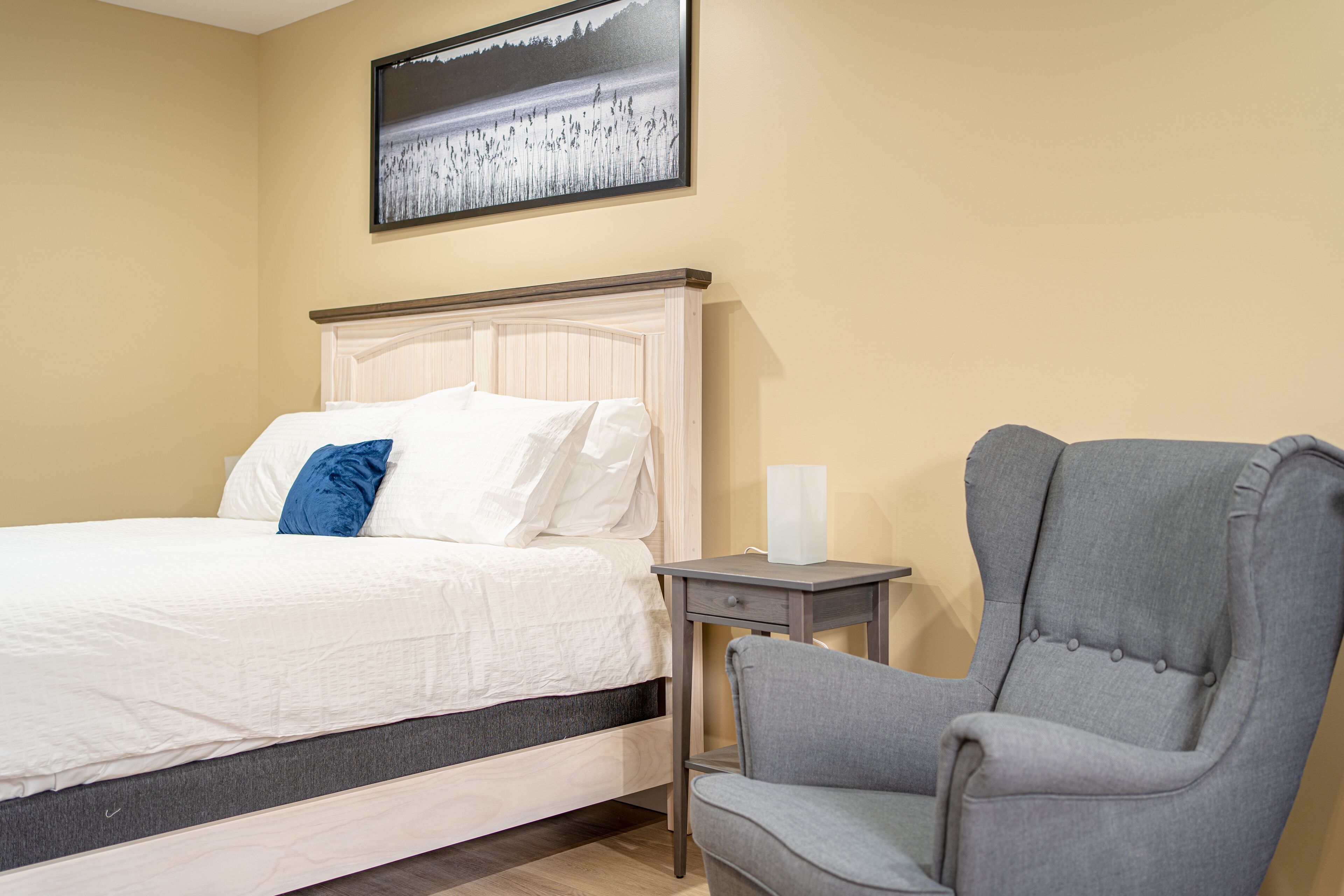 A neatly made bed with white and blue pillows, a grey upholstered armchair, and a small bedside table with a white lamp.