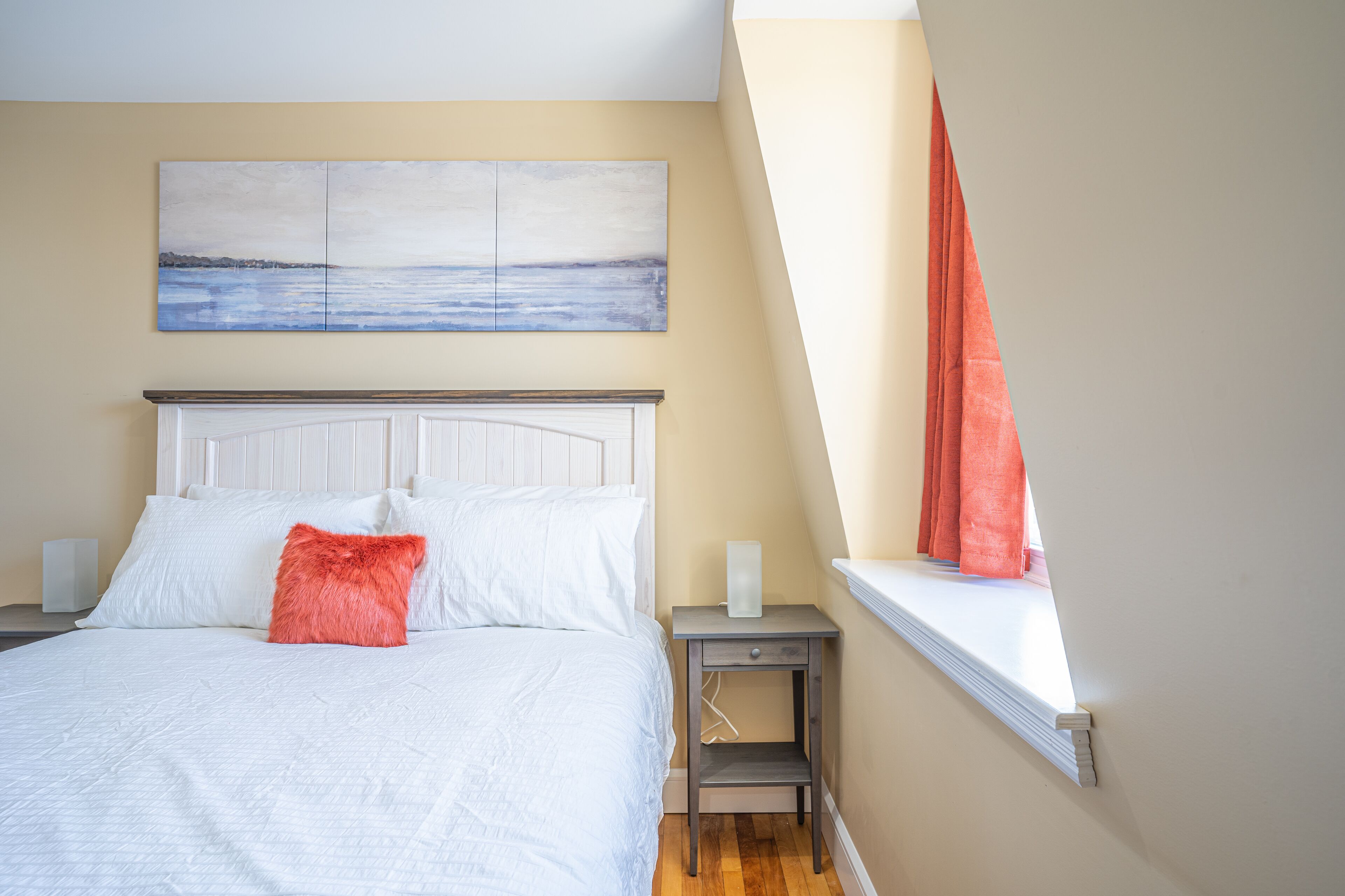 A bedroom with a bed, a nightstand, a window with curtains, and a painting on the wall.