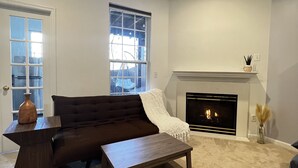 A living room with a dark brown sofa, a wooden coffee table, a fireplace, and a large window. - Woodland Park Studio (Herndon)