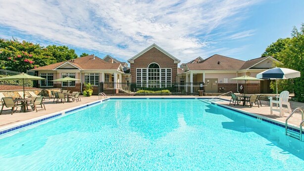 A swimming pool with lounge chairs and umbrellas, surrounded by brick buildings with a central arched entrance. - Woodland Park Studio (Herndon)