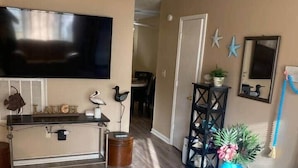 A living room with a flat-screen TV, a console table, a blue-striped rug, a dark wooden floor, a door, a small plant, and wall decorations. - Pink Flamingo-Pet-Friendly + Walk to Beach & Dining, Fenced courtyard (North Myrtle Beach)