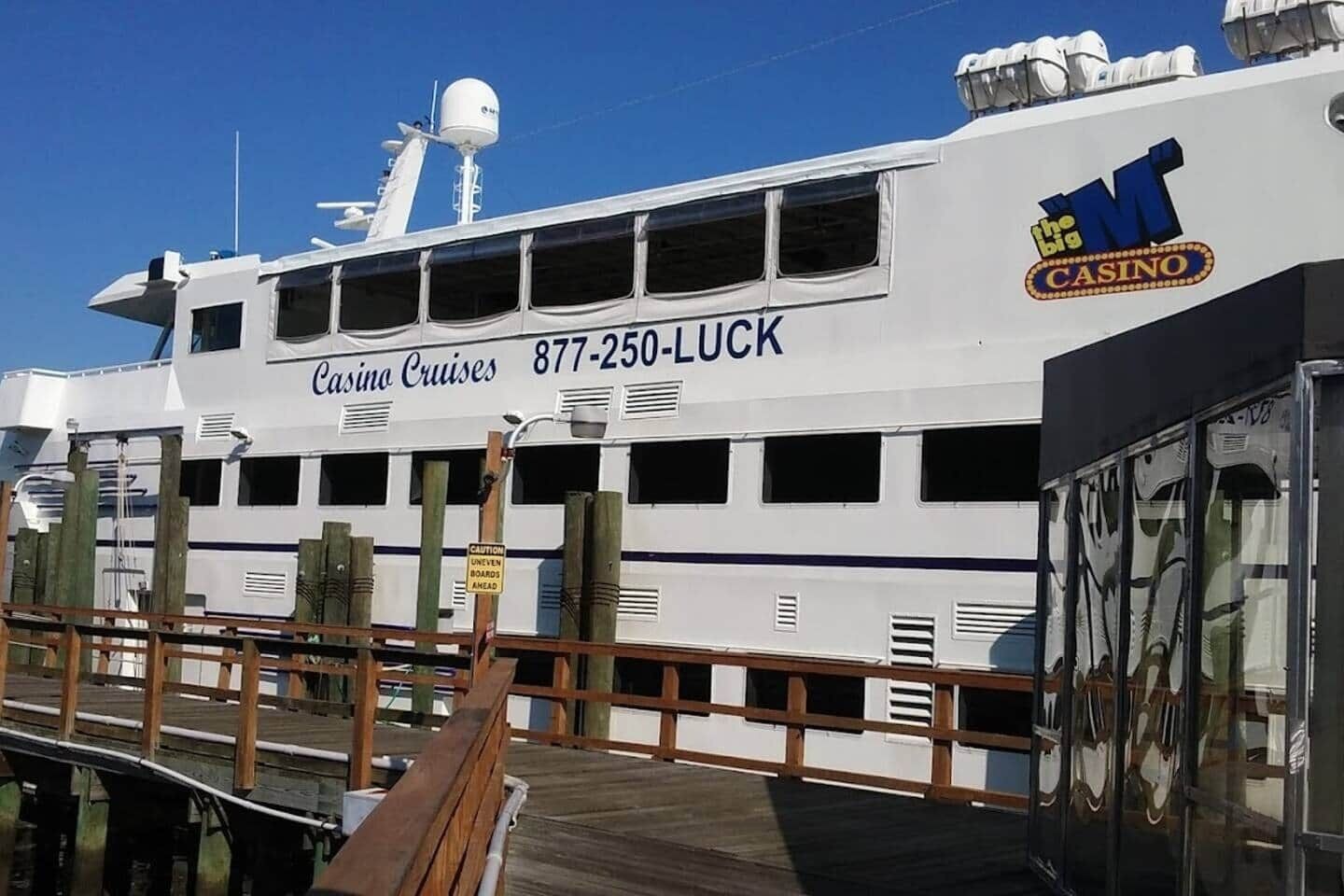 Un gran barco crucero blanco con la marca 'Casino Cruises' y 'The Big Casino', un número de teléfono y una cubierta de madera.