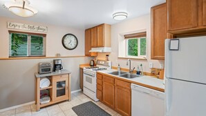 A kitchen with wooden cabinets, a stove, a sink, and a refrigerator. There is a clock on the wall and a sign that says "Enjoy the little things." - Wind Song Retreat on the Anchorage Hillside! (Anchorage)
