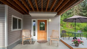 A covered porch with two wooden chairs, a wooden door, and a patio table with an umbrella. - Wind Song Retreat on the Anchorage Hillside! (Anchorage)
