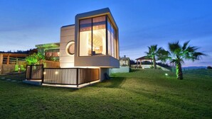 Una casa moderna a un piano con un'ampia vetrata, immersa in un prato curato e circondata da palme.