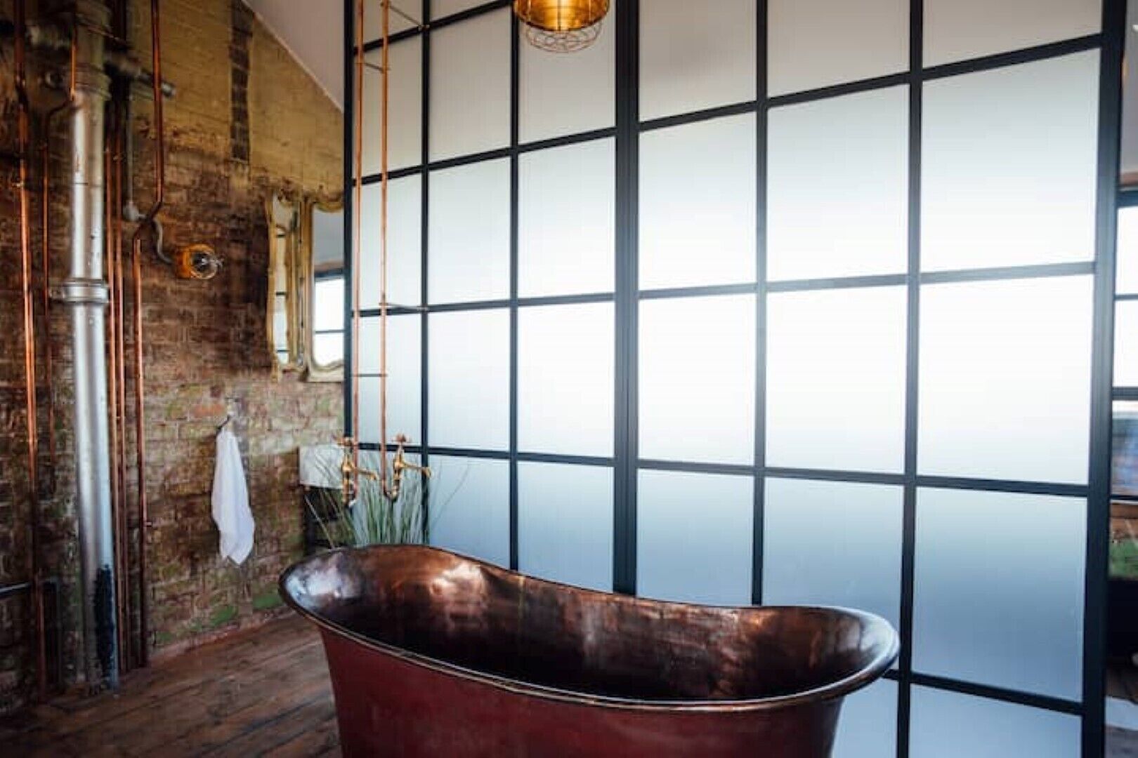 A bathroom with a large copper bathtub, exposed brick walls, and a grid-patterned window.