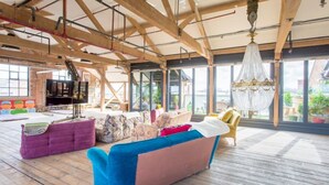 A spacious living area with a high ceiling, wooden beams, and large windows offering a view of the outdoors.