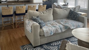 Ein gemütliches Wohnzimmer mit grauem Sofa, Korbsesseln, einem Couchtisch und einer Küchenzeile mit Holzschränken und einem Kühlschrank.