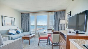 A hotel room with a sofa, a dining table with chairs, a TV, and a view of the sea through large windows. - Thames Street - Newport Harbor 1 Bedroom (Newport)