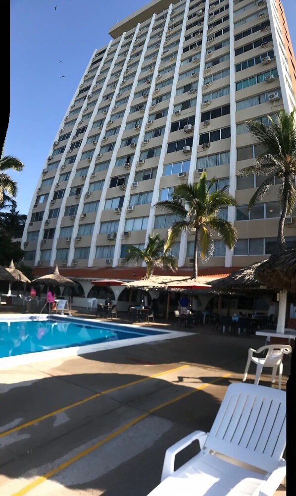 A high-rise hotel with a swimming pool, palm trees, and outdoor seating.