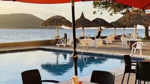 Un espace aménagé au bord de la piscine, avec une table et des chaises sous un parasol rouge, entouré de chaises longues et offrant une vue sur la mer.