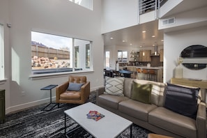 Una sala de estar moderna con un sofá, un sillón y una mesa de centro. Un ventanal ofrece vistas a un patio trasero con rocas y una valla de madera. Se aprecia una cocina al fondo con taburetes de bar.