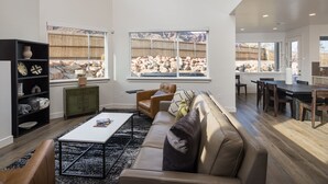 Un salón amplio con un sofá de cuero, una mesa de centro y una estantería. Ventanas grandes ofrecen vistas a un paisaje rocoso.