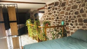 Quarto com parede de pedra, cama com colcha azul, treliça de madeira com plantas e prateleira de metal com cestos trançados.