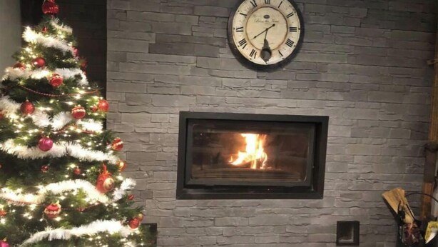 Árvore de Natal com enfeites vermelhos e luzes, lareira com fogo visível e relógio de parede acima da lareira.