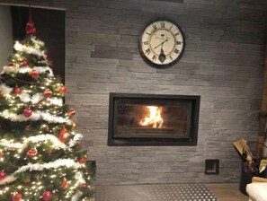 Un sapin de Noël décoré d’ornements rouges et de guirlandes lumineuses, une cheminée avec un feu visible et une horloge murale au-dessus de la cheminée.