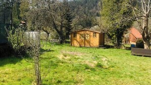 Uma pequena cabana de madeira numa área gramada, com árvores e uma cerca ao fundo.