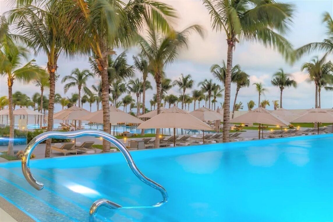 A swimming pool with a curved edge, surrounded by palm trees and lounge chairs under umbrellas.