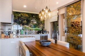 A modern kitchen with a wooden dining table, a large island with a built-in stove, and a view of a lush garden through a large window.