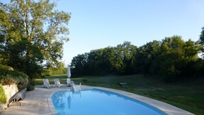 Ein Schwimmbecken mit einer gefliesten Terrasse, umgeben von einer Rasenfläche und Bäumen.