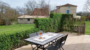 Eine Terrasse mit einem gefliesten Tisch und Stühlen, einer Weinflasche und Blick auf ein Haus aus Stein mit einem blauen Fenster.