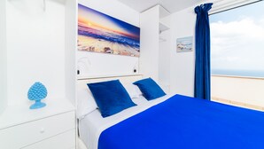 A bedroom with a bed, a painting of a beach sunset, a window with a view of the sea, and a blue curtain.