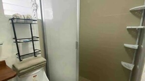 A bathroom with a toilet, a shower enclosure, and a shelf with toiletries. - Depas Tramonto (Mazatlán)