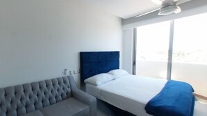 A modern hotel room with a grey sofa, a bed with a blue headboard, and a large window providing a view. - Depas Tramonto (Mazatlán)