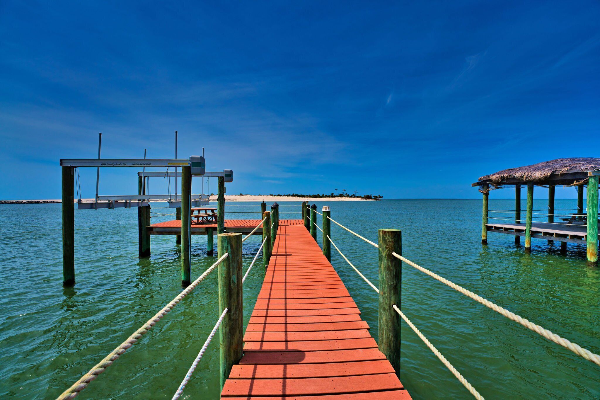 A wooden pier extending into calm waters, with a thatched-roof structure on the right and a metal railing on the left.