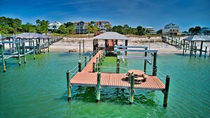 Une jetée dotée de passerelles en bois et d’un espace de repos abrité, qui mène à une plage où l’on aperçoit des maisons au loin.