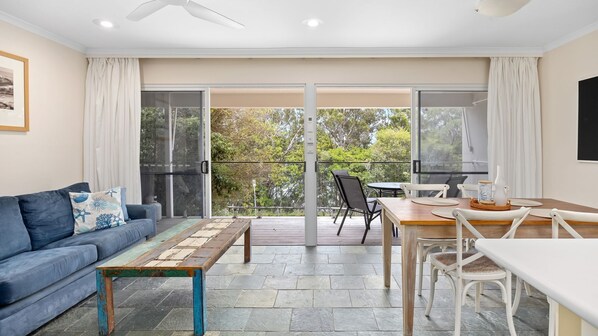 A modern living room with a blue sofa, a wooden coffee table, a dining table with chairs, and a balcony with glass doors. - Alderly Terrace 1 Noosa Heads (Noosa Heads)