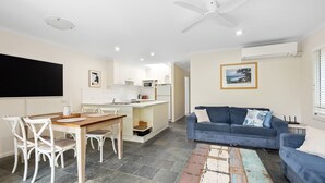 Una sala moderna con un sofá azul, un comedor con mesa de madera y sillas blancas, y una cocina con armarios blancos y un ventilador de techo.