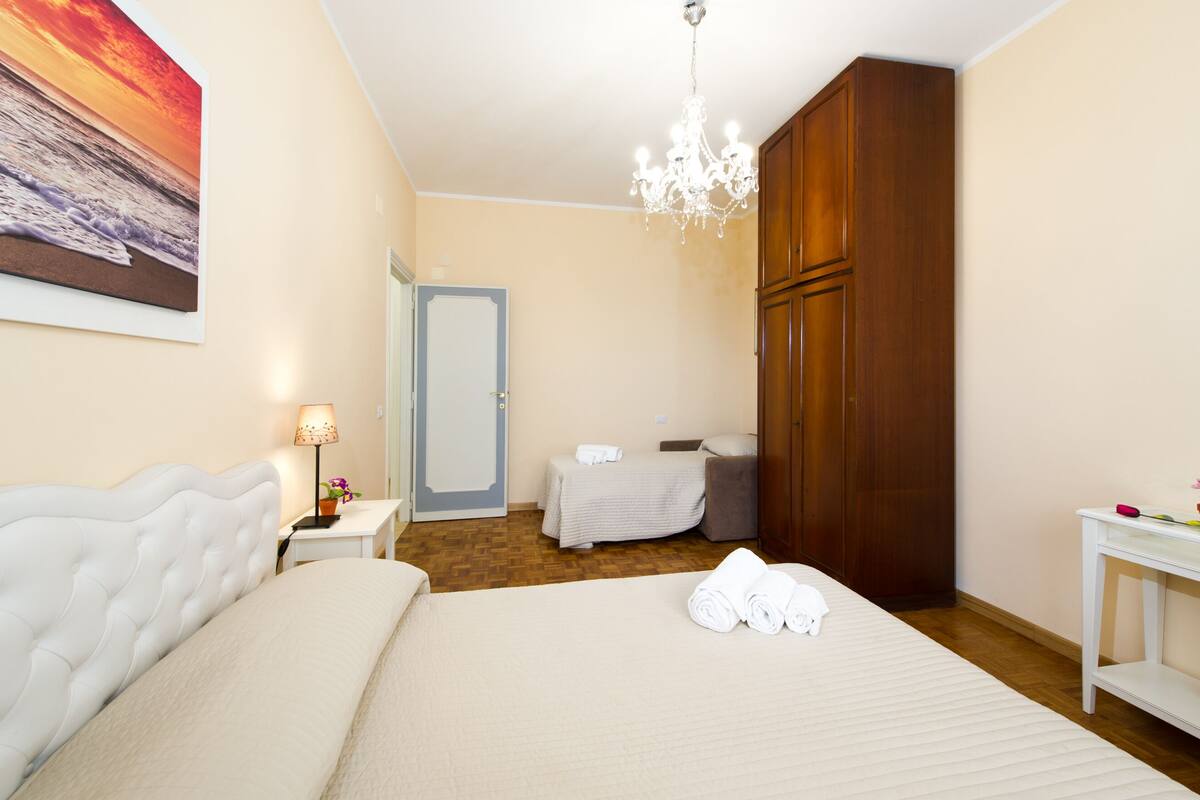 Quarto de hotel com cama, mesa de cabeceira, luminária, guarda-roupa e pintura de um pôr do sol sobre o oceano.