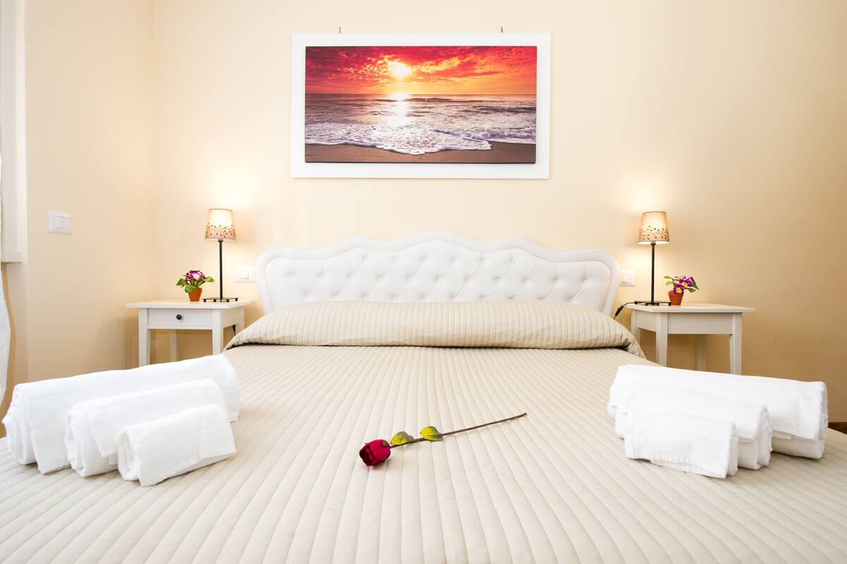 Quarto de hotel com uma cama grande, duas mesas de cabeceira com abajures e um quadro com uma paisagem de pôr do sol sobre o oceano.