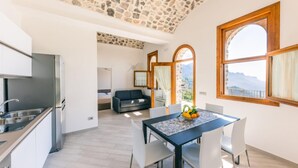 A modern kitchen with a built-in stove, a dining area with a table and chairs, and a view of mountains through large windows. - Casa Meraviglia Ravello (RAVELLO)